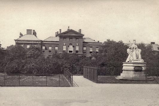 Black and white photograph of Kensington Palace, England, with a marble statue in the foreground and some shrubs that lie before it, taken in the late 19th century.