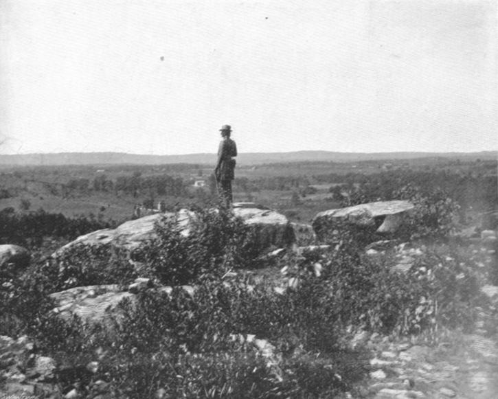 Black and white photograph of a statue of a Civil War Soldier in uniform on a hill overlooking lower lands.